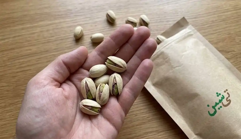 دست در حال گرفتن پسته اکبری خام با پوسته باز طبیعی و مغز سبز روی میز چوبی تی شین